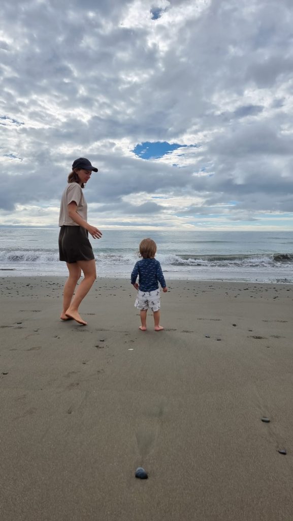 Alannah walking barefoot on a sandy beach beside her toddler, both facing the waves under a cloudy sky